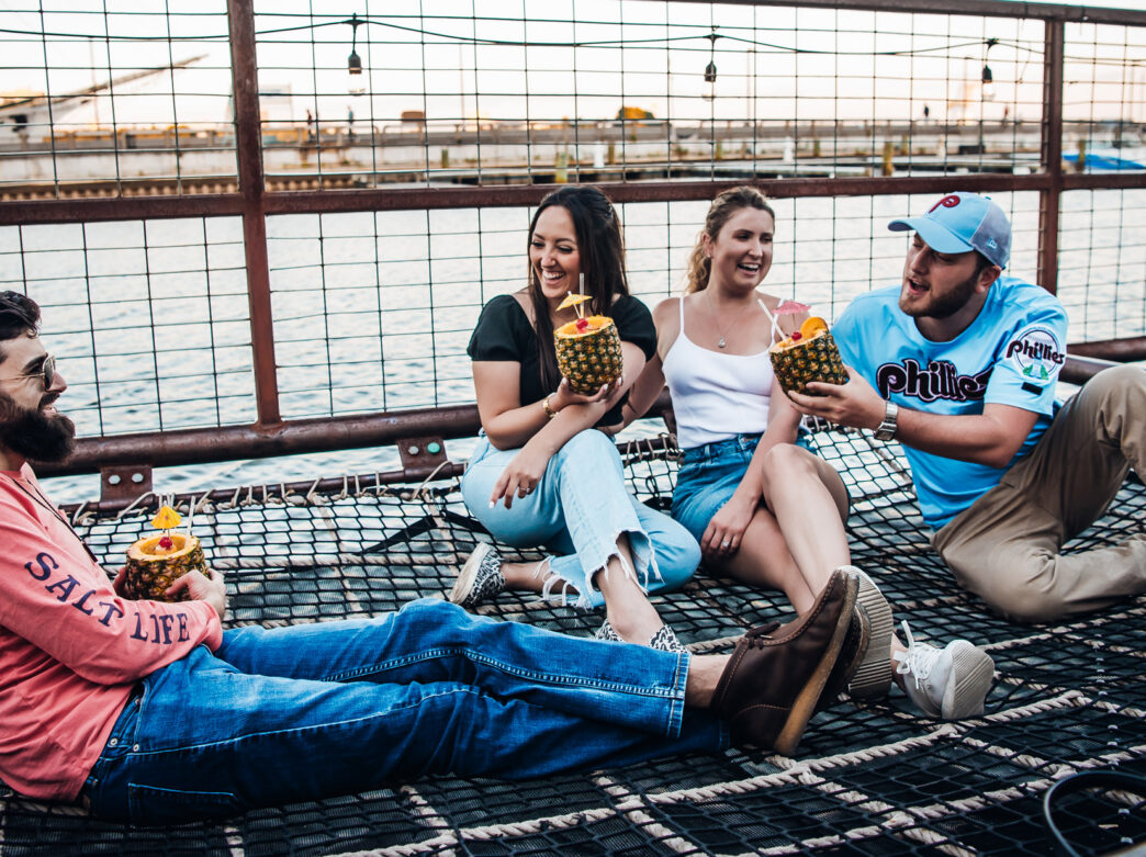 Four friends relax together on a netted lounge area at Spruce Street Harbor Park in Philadelphia, laughing and holding tropical pineapple drinks beside the Delaware River at sunset. String lights hang overhead while boats and the waterfront create a lively summer atmosphere in the background.