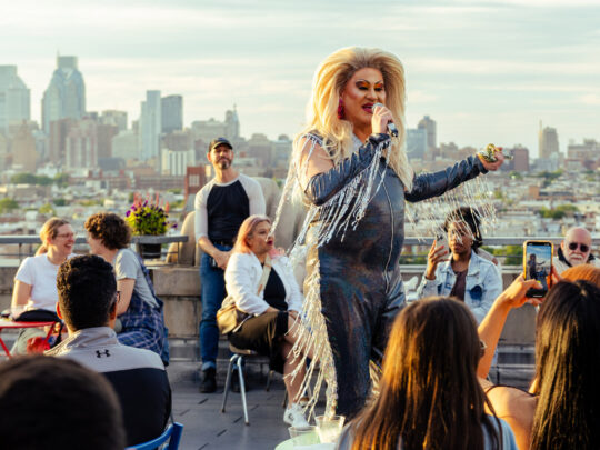 A drag performer in a black outfit with sparkling fringe speaks into a microphone at Bok Bar with a view of the Philadelphia skyline in the background.