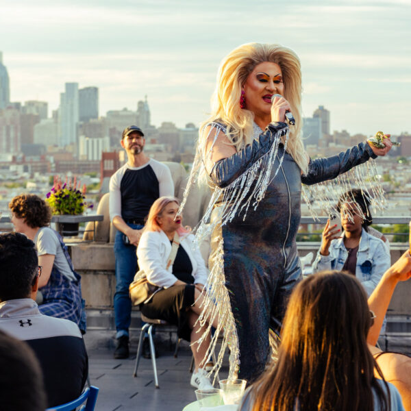 A drag performer in a black outfit with sparkling fringe speaks into a microphone at Bok Bar with a view of the Philadelphia skyline in the background.