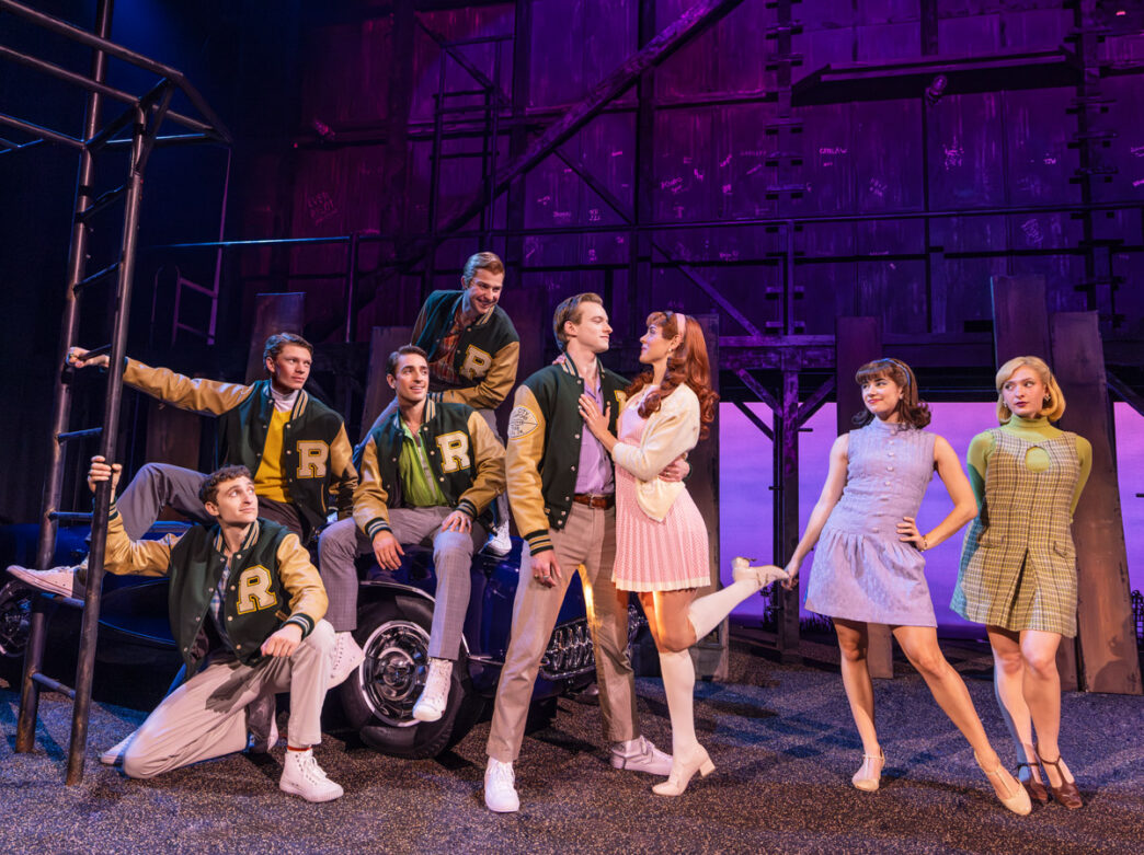 Cast members perform a scene from The Outsiders onstage, wearing 1960s-style costumes and letterman jackets around a car beneath dramatic purple theater lighting.