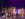 Cast members perform a scene from The Outsiders onstage, wearing 1960s-style costumes and letterman jackets around a car beneath dramatic purple theater lighting.