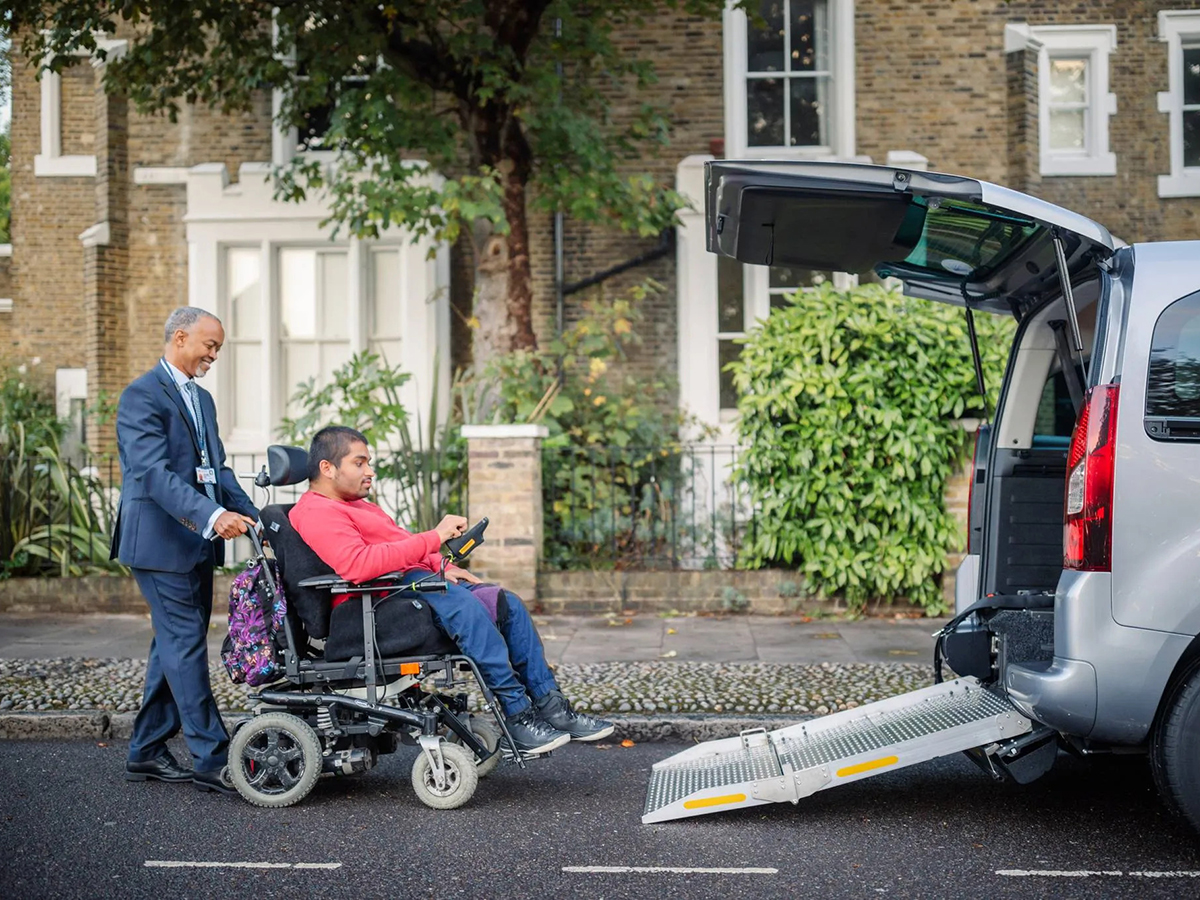 A person wearing a navy suit pushes a person in a wheelchair up a ramp into a wheelchair-accessible vehicle.
