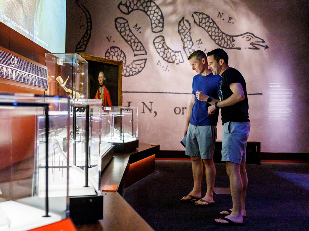 Two visitors examine historical artifacts in glass cases at the Museum of the American Revolution, with a large political cartoon displayed on the wall behind them.